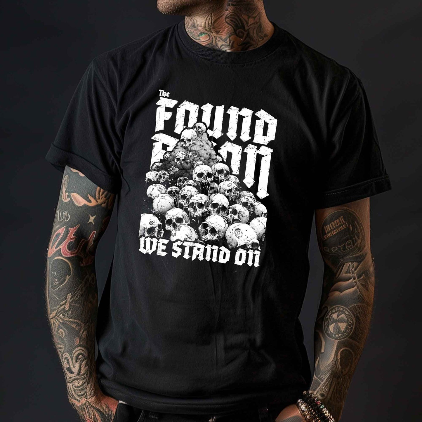 T-Shirt mit einem Berg aus Totenköpfen und dem Slogan 'The foundation we stand on'. Das Design ist Weiß auf schwarzem/dunkeln Stoff und thematisiert die brutalen Hintergründe von Landbesitz und Eroberung.
