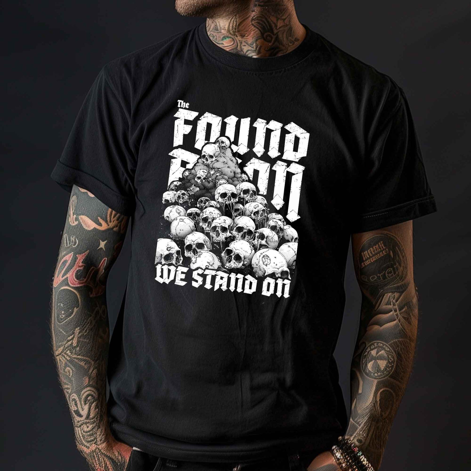 T-Shirt mit einem Berg aus Totenköpfen und dem Slogan 'The foundation we stand on'. Das Design ist Weiß auf schwarzem/dunkeln Stoff und thematisiert die brutalen Hintergründe von Landbesitz und Eroberung.