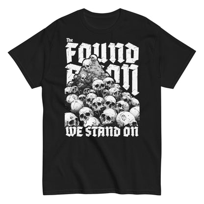 T-Shirt mit einem Berg aus Totenköpfen und dem Slogan 'The foundation we stand on'. Das Design ist Weiß auf schwarzem/dunkeln Stoff und thematisiert die brutalen Hintergründe von Landbesitz und Eroberung.